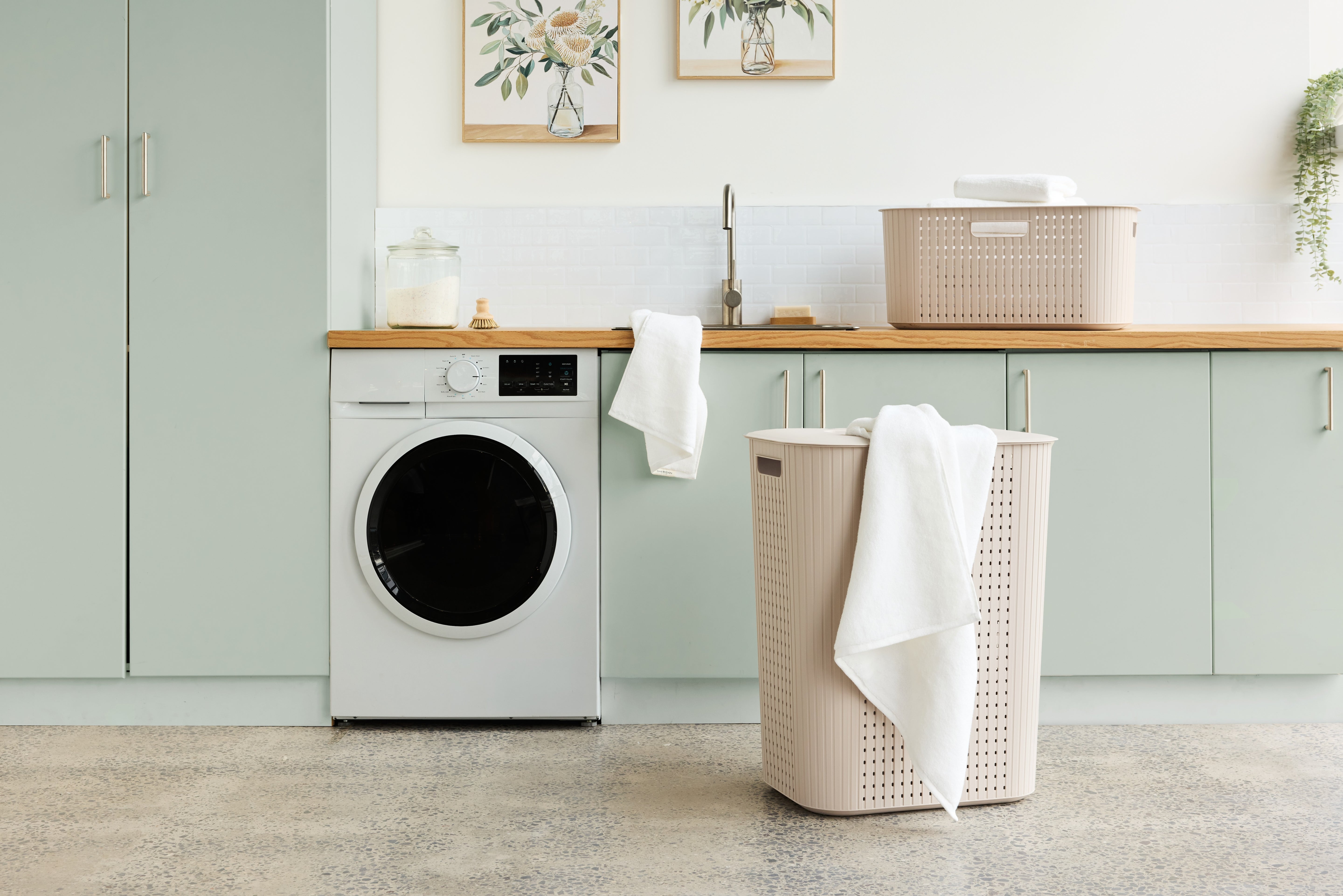 A modern laundry room scene with a white front-loading washing machine, pale green cabinets, a wooden countertop, and a beige laundry hamper draped with a white towel in the foreground.