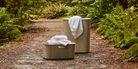 Two metallic beige perforated laundry baskets with white towels, placed on a dirt path surrounded by lush green ferns.