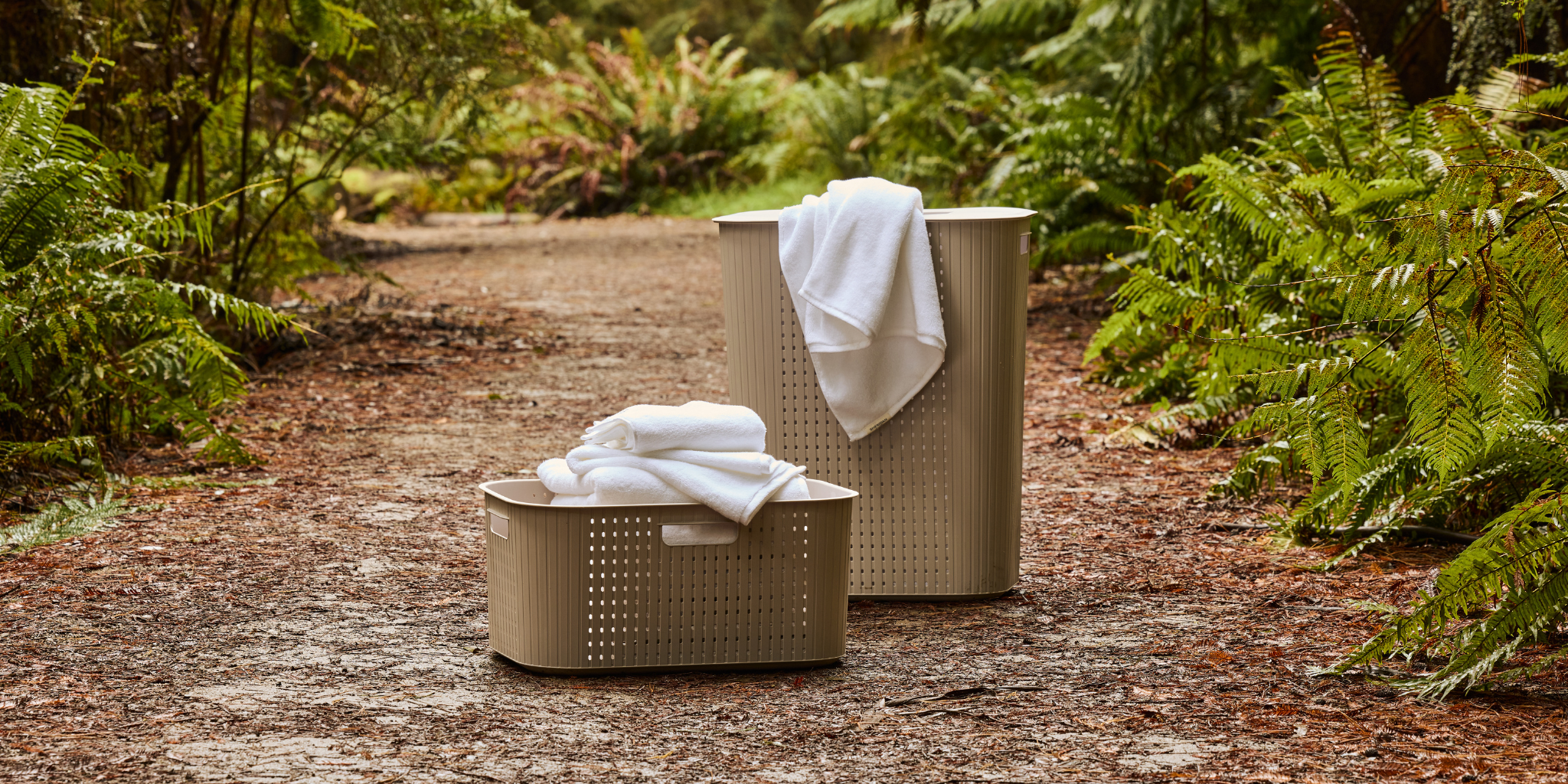 Two metallic beige perforated laundry baskets with white towels, placed on a dirt path surrounded by lush green ferns.